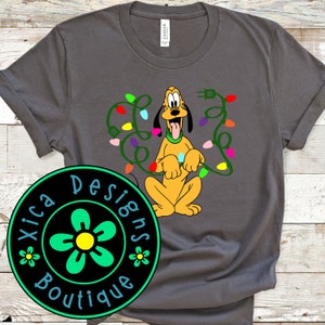 May include: A grey t-shirt featuring a cartoon image of Pluto the dog entangled in Christmas lights. The shirt has a green circle logo with a flower and the text "Xica Designs Boutique".