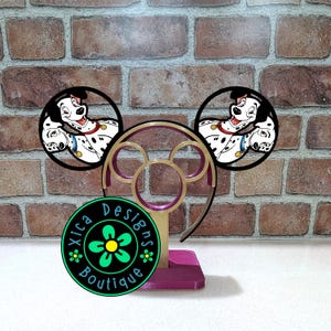 May include: A gold and magenta plastic display stand for headbands featuring a circular base and a central frame. Two circular black frames are attached to the top, each displaying a cartoon Dalmatian dog. A circular logo reads Xica Designs Boutique.