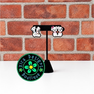 May include: Two pairs of dalmatian dog earrings displayed on a black T-shaped stand. The left pair features two dalmatians with blue and red collars, while the right pair shows two dalmatians with a pink bow and a red collar. A circular logo reads Xica Designs Boutique.