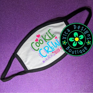 May include: White face mask with black trim featuring the text "Cookie Crew" in green, blue, and pink with hearts and a small arrow pointing to the right. The text "Troup #" is below the arrow. A black circle with a green flower and the text "Xica Designs Boutique" is in the background.