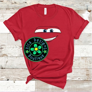 Pu&ograve; includere: T-shirt rossa con il volto di un'auto dei cartoni animati, con occhi blu e bocca sorridente. Sul davanti, un logo circolare con la scritta "Xica Designs Boutique" e un motivo floreale.