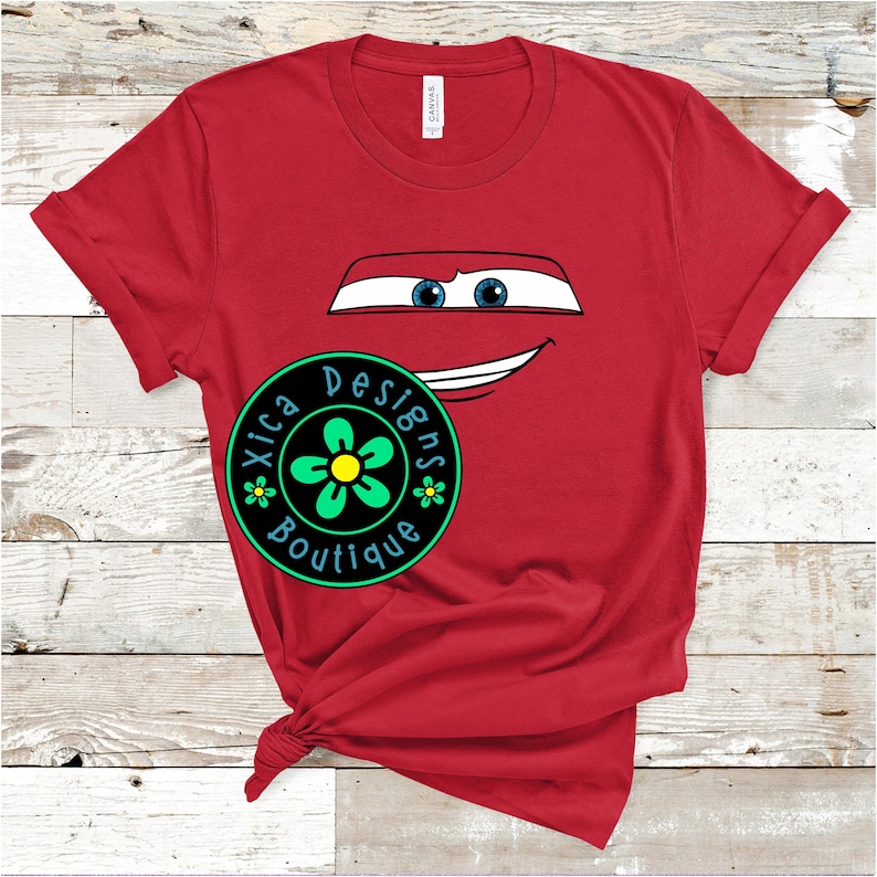 Pu&ograve; includere: T-shirt rossa con il volto di un'auto dei cartoni animati, con occhi blu e bocca sorridente. Sul davanti, un logo circolare con la scritta "Xica Designs Boutique" e un motivo floreale.