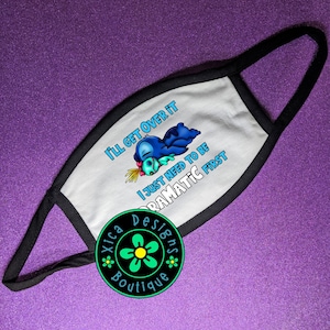 K&ouml;nnte beinhalten: Wei&szlig;e Gesichtsmaske mit schwarzem Rand und Ohrenschlaufen. Die Maske zeigt eine Zeichentrickfigur und den Text "I'LL GET OVER IT I JUST NEED TO BE DRAMATIC FIRST." Ein kreisf&ouml;rmiges Logo befindet sich in der unteren linken Ecke.