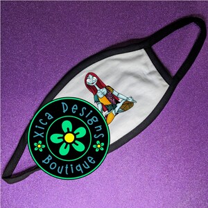 May include: White face mask with a black border. The mask features a colorful illustration of a character from the Nightmare Before Christmas movie. The mask is decorated with a green flower and the text "Xica Designs Boutique".