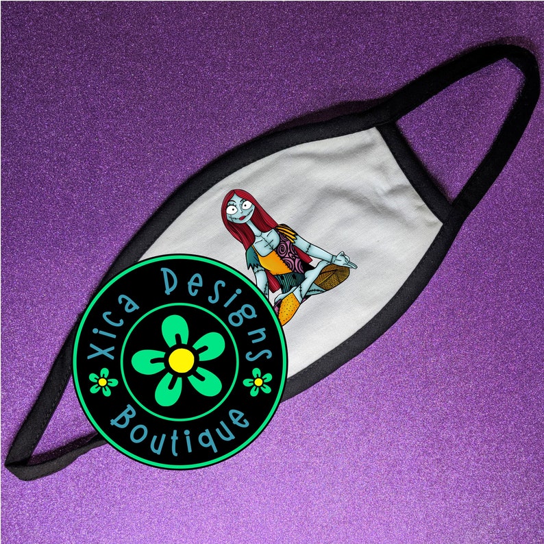 May include: White face mask with a black border. The mask features a colorful illustration of a character from the Nightmare Before Christmas movie. The mask is decorated with a green flower and the text "Xica Designs Boutique".