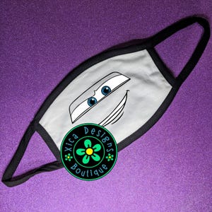 Pu&ograve; includere: Mascherina grigia con un design a forma di volto di auto dei cartoni animati, bordi neri e passanti neri. La mascherina ha occhi blu e una bocca sorridente. Un logo circolare con la scritta "Xica Designs Boutique" &egrave; sulla mascherina.