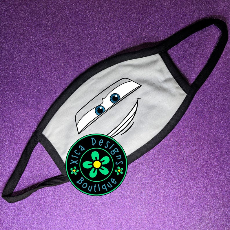 Pu&ograve; includere: Mascherina grigia con un design a forma di volto di auto dei cartoni animati, bordi neri e passanti neri. La mascherina ha occhi blu e una bocca sorridente. Un logo circolare con la scritta "Xica Designs Boutique" &egrave; sulla mascherina.
