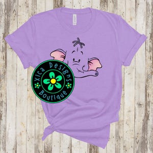 May include: A light purple t-shirt featuring a cartoon elephant face with pink ears and a trunk. The shirt has a round logo with the text "Xica Designs Boutique" and a flower design. The shirt is made of a soft material.