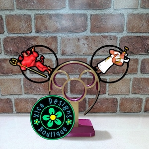 May include: A gold-colored display stand for headbands featuring a circular frame with two attached black circular cutouts. The left cutout displays a red devil character with a trident, and the right shows a white-robed angel. A circular logo reads Xica Designs Boutique.