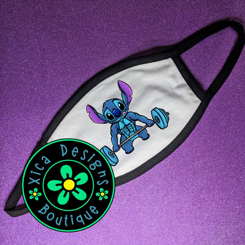 Pode incluir: M&aacute;scara facial branca com acabamento e presilhas pretas. A m&aacute;scara apresenta uma ilustra&ccedil;&atilde;o de desenho animado de um alien&iacute;gena azul levantando pesos. Um logotipo circular com o texto "Xica Designs Boutique" est&aacute; no canto inferior esquerdo.