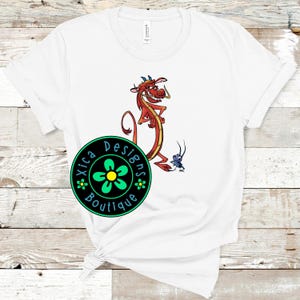 May include: White t-shirt featuring a cartoon dragon in red and yellow with a small blue insect. The shirt also has a circular logo with the text "Xica Designs Boutique" around a green flower.