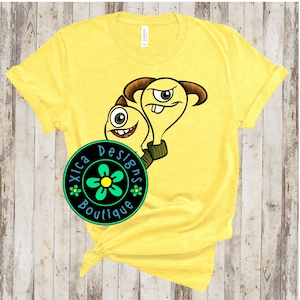 May include: Yellow t-shirt with a graphic of two cartoon monsters. The monsters are green and yellow with large eyes and toothy grins. The text "Xica Designs Boutique" is printed in a circle on the shirt.