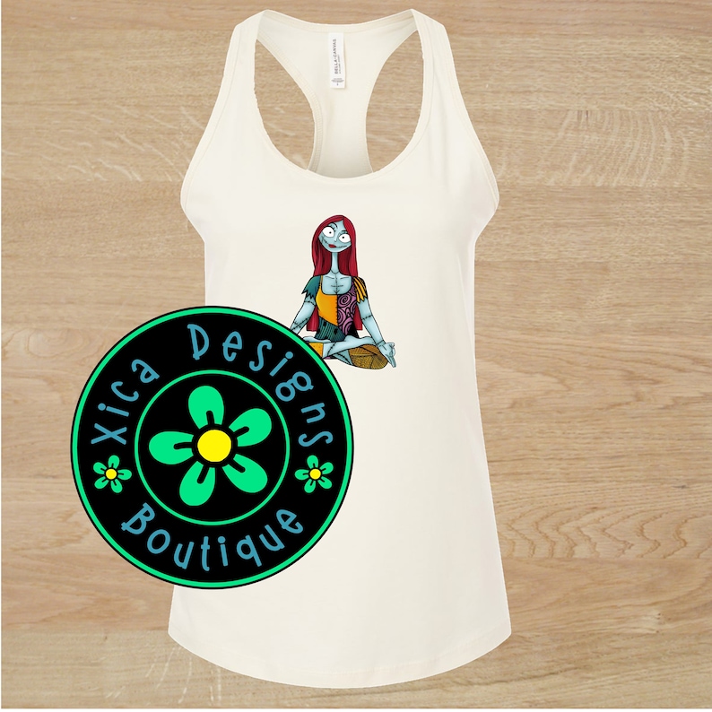 May include: White tank top with a graphic of a red-haired woman in a yoga pose. The graphic is of a character from the movie "The Nightmare Before Christmas". The tank top has a black circle with a green flower and the text "Xica Designs Boutique" on it.
