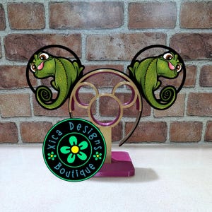 May include: A decorative stand featuring a Mickey Mouse ear design with two green chameleon figures. The stand has a purple base and gold accents. A circular logo with the text "Xica Designs Boutique" is visible.