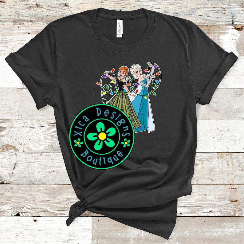 Peut inclure: T-shirt noir avec un graphisme de deux femmes anim&eacute;es tenant des guirlandes lumineuses. Le design comprend le texte "Xica Designs Boutique" dans un logo circulaire avec une fleur. Le t-shirt est fait d'une mati&egrave;re douce.