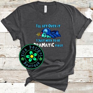 K&ouml;nnte beinhalten: Dunkelgraues T-Shirt mit einer Cartoonfigur in Blau und T&uuml;rkis, mit dem Text "I'll Get Over It, I Just Need To Be Dramatic First." Ein gr&uuml;nes und schwarzes kreisf&ouml;rmiges Logo mit dem Text "Xica Designs Boutique" befindet sich unten.