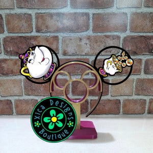 May include: A decorative headband featuring a Mickey Mouse ear design with Beauty and the Beast character accents. The headband has a purple and gold color scheme, with a black and green circular logo that reads "Xica Designs Boutique".