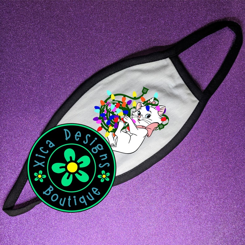 Peut inclure: Masque facial blanc avec une bordure noire. Le masque pr&eacute;sente une image de dessin anim&eacute; d'un chat blanc emm&ecirc;l&eacute; dans des lumi&egrave;res de No&euml;l color&eacute;es. Le texte "Xica Designs Boutique" est imprim&eacute; en cercle sur le masque.