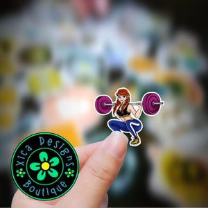 May include: A sticker featuring a cartoon woman with red hair lifting a barbell. She is wearing a blue and black workout outfit. The sticker is on a hand, and the background is blurry.