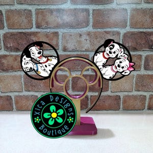 May include: A gold and magenta plastic display stand for headbands featuring two circular frames with illustrations of Dalmatian dogs. The left circle shows two adult Dalmatians, and the right circle shows two puppies. A circular logo reads Xica Designs Boutique.