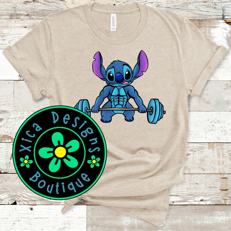 Pode incluir: Camiseta bege com um personagem de desenho animado levantando pesos. O personagem azul tem orelhas grandes e segura uma barra azul. Um logotipo circular com o texto "Xica Designs Boutique" est&aacute; no canto inferior esquerdo.