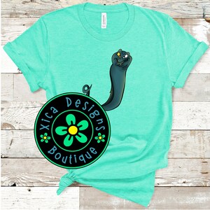 Puede incluir: Una camiseta turquesa con un logotipo circular negro que dice "Xica Designs Boutique" con una flor verde en el centro. Un serpiente marina de dibujos animados asoma por detr&aacute;s del logotipo.