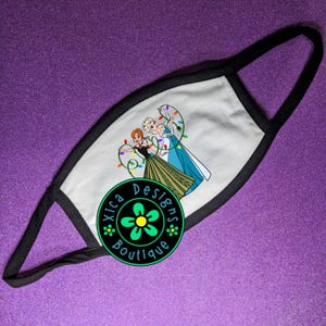 Peut inclure: Un masque facial blanc avec des bordures et des boucles noires. Le masque pr&eacute;sente une illustration de dessin anim&eacute; de deux personnages tenant des guirlandes lumineuses. Le masque porte un logo circulaire qui dit "Xica Designs Boutique".