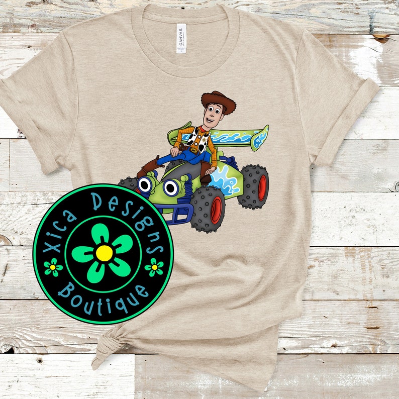 May include: A beige t-shirt with a green, blue, and black cartoon image of Woody from Toy Story driving a green race car. The image is surrounded by a green circle with a black background and white text that reads "Xica Designs Boutique".