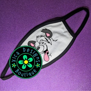 May include: White face mask with a black border. The mask features a cartoon dog with a pink tongue sticking out. The mask has a black and green circle with a white flower and the text "Xica Designs Boutique" on it.