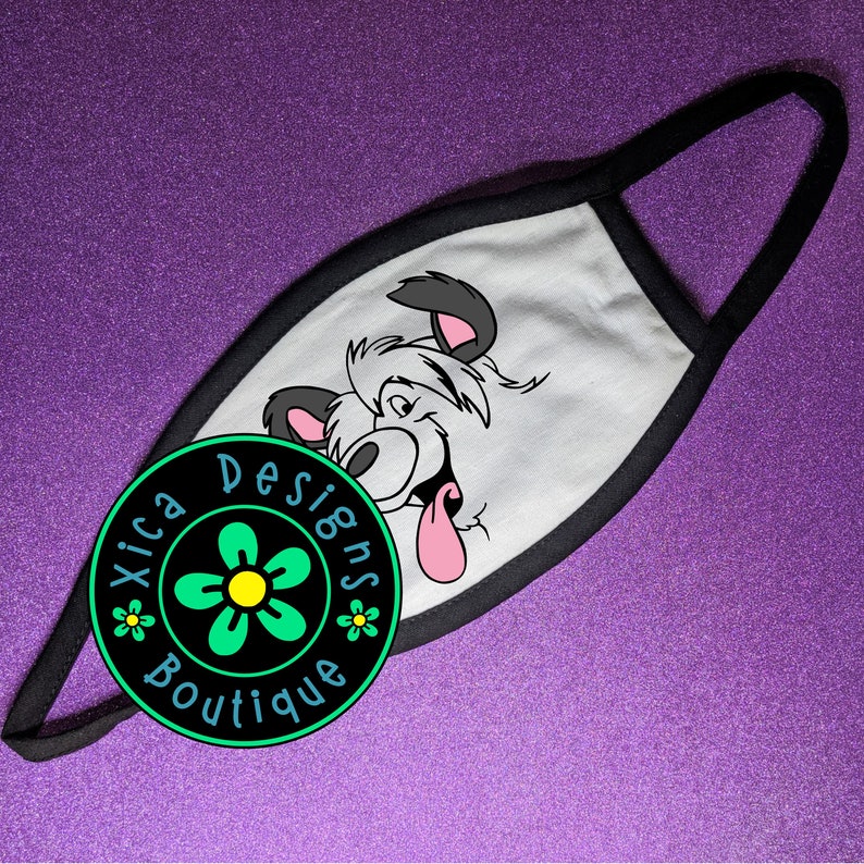 May include: White face mask with a black border. The mask features a cartoon dog with a pink tongue sticking out. The mask has a black and green circle with a white flower and the text "Xica Designs Boutique" on it.