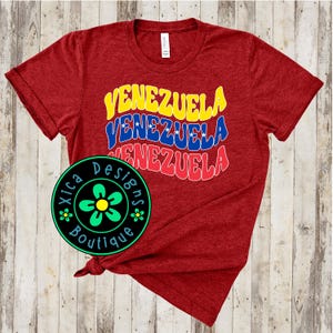 May include: Red t-shirt with the word "Venezuela" repeated three times in a retro font with yellow, blue, and red colours. The shirt is tied in a knot and a small green flower logo is visible in the bottom left corner.