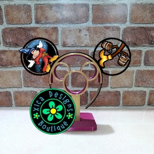 May include: A decorative headband with Mickey Mouse ears. The ears feature circular designs with images of Mickey Mouse and a broom carrying buckets. The headband has a gold and purple color scheme, and the base is purple. The logo for Xica Designs Boutique is also visible.