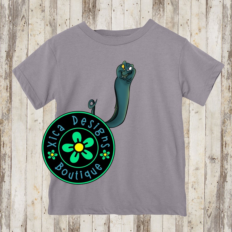 K&ouml;nnte beinhalten: Ein graues T-Shirt mit einem gr&uuml;nen und gelben Blumendesign und dem Text "Xica Designs Boutique" in einem schwarzen Kreis. Ein Cartoon-Aal mit einem schelmischen Ausdruck ist auf das Shirt gedruckt.