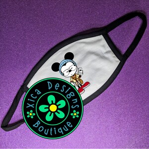 May include: White face mask with a black border. The mask features a cartoon illustration of Mickey Mouse wearing glasses and a blue hat. The mask is decorated with a black circle with the text "Xica Designs Boutique" and a green flower.