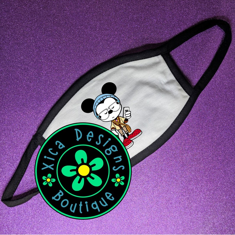 May include: White face mask with a black border. The mask features a cartoon illustration of Mickey Mouse wearing glasses and a blue hat. The mask is decorated with a black circle with the text "Xica Designs Boutique" and a green flower.