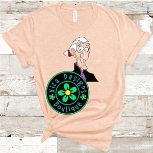 May include: A peach-coloured t-shirt featuring a cartoon image of a man with a white beard and a purple shirt. The t-shirt also has a black circle with a green flower and the text "Xica Designs Boutique" in white.