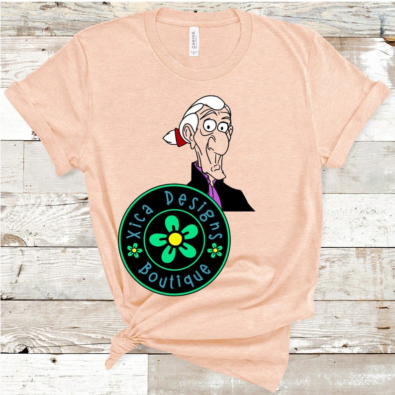 May include: A peach-coloured t-shirt featuring a cartoon image of a man with a white beard and a purple shirt. The t-shirt also has a black circle with a green flower and the text "Xica Designs Boutique" in white.