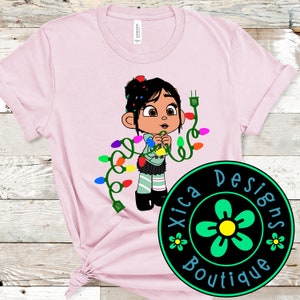 May include: Pink t-shirt featuring a cartoon character, Vanellope von Schweetz from Wreck-It Ralph, holding Christmas lights. The shirt has a black and green circle with a flower design and the text "Xica Designs Boutique".
