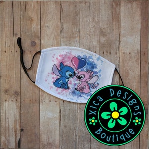 May include: A white face mask with a blue and pink cartoon couple kissing. The background is a watercolor design with blue and pink splatters. The mask has a black elastic strap. The mask is labeled "Xica Designs Boutique".