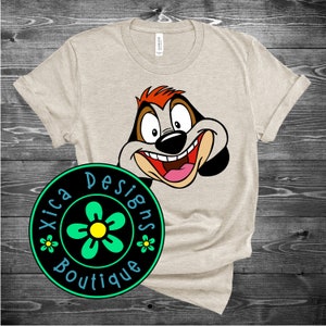 May include: A beige t-shirt with a cartoon image of a dog's face. The dog has red hair, a black nose, and a big smile. The t-shirt has a black circle with a green flower and the text "Xica Designs Boutique" in white.