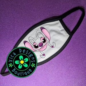 Puede incluir: M&aacute;scara facial blanca con borde negro. La m&aacute;scara presenta una imagen de dibujos animados de un extraterrestre gris con orejas rosas y una nariz morada. El texto "Xica Designs Boutique" est&aacute; impreso en un c&iacute;rculo negro en la esquina inferior izquierda de la imagen.