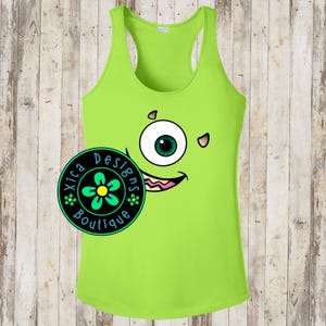 May include: Lime green racerback tank top featuring a cartoon monster face design. The monster has one large eye with a green iris, a pink mouth, and small details. The tank top has the text "Xica Designs Boutique" in a circular logo.