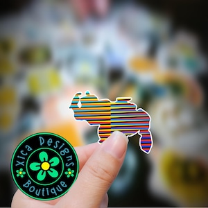 May include: A colorful sticker of the outline of Venezuela with a striped pattern in yellow, blue, red, and black. The sticker is being held by a hand. The sticker is labeled "Xica Designs Boutique".
