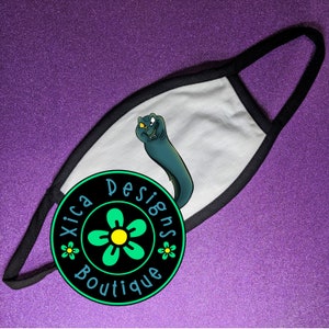 K&ouml;nnte beinhalten: Wei&szlig;e Gesichtsmaske mit schwarzem Rand und einem Cartoon-Bild eines gr&uuml;nen Seeschlangens. Die Maske hat ein schwarzes und gr&uuml;nes Blumenlogo mit dem Text "Xica Designs Boutique".