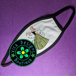 Pu&ograve; includere: Maschera facciale bianca con bordo nero. La maschera presenta un'immagine di cartone animato di una donna con un vestito verde con luci colorate avvolte intorno a lei. Il testo "Xica Designs Boutique" &egrave; stampato su un cerchio verde con un fiore giallo.