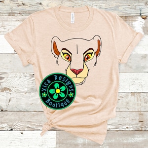 May include: A beige t-shirt with a cartoon lion face graphic. The lion has orange fur, yellow eyes, and a pink nose. The shirt also has a small green flower logo with the text "Xica Designs Boutique".