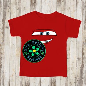 Pu&ograve; includere: Una maglietta rossa con un disegno di un volto di auto dei cartoni animati, con occhi blu e una bocca sorridente. Un logo circolare con la scritta "Xica Designs Boutique" e un disegno floreale si trova in basso a sinistra. La maglietta &egrave; in materiale morbido.