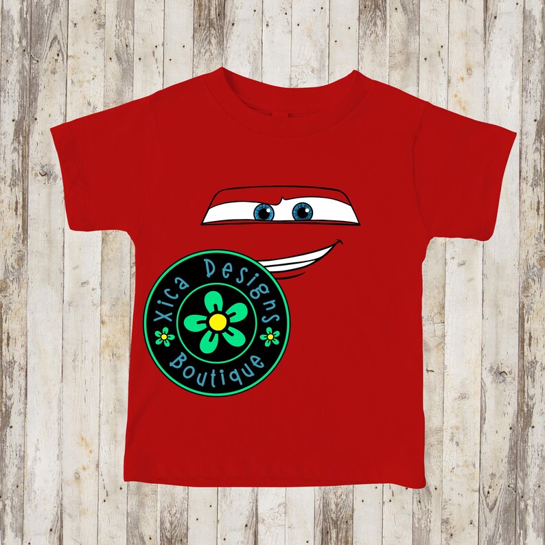 Pu&ograve; includere: Una maglietta rossa con un disegno di un volto di auto dei cartoni animati, con occhi blu e una bocca sorridente. Un logo circolare con la scritta "Xica Designs Boutique" e un disegno floreale si trova in basso a sinistra. La maglietta &egrave; in materiale morbido.