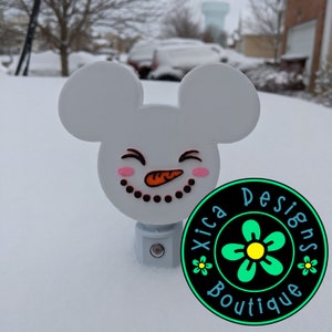 May include: White snowman figurine with Mickey Mouse ears, a carrot nose, and black eyes. The snowman is smiling and has pink cheeks. The figurine is sitting on a white surface. The text "Xica Designs Boutique" is visible in the background.