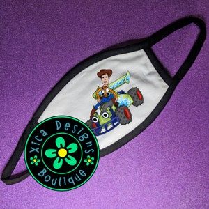 May include: White face mask with a black border. The mask features a cartoon image of Woody from Toy Story driving a green dune buggy. The mask has a black circle with a green flower and the text "Xica Designs Boutique" printed on it.
